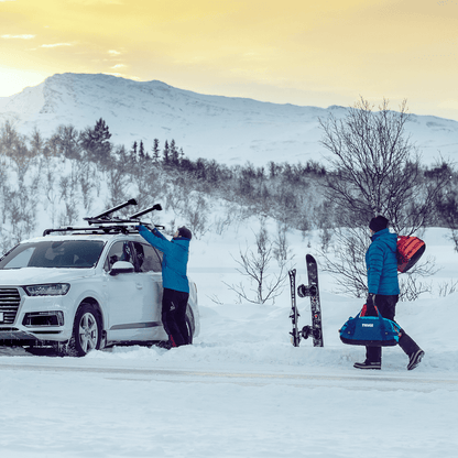 A man is unloading skis from his car roof with a Thule SnowPack Extender while another man is walking towards him with Thule Chasm bags.