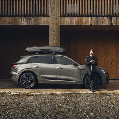 A man leans against a vehicle with a Thule Motion 3 roof box parked next to a wooden wall.