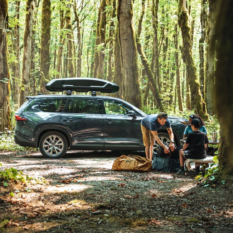 In a forest setting, a man leans over various Thule bags while a woman and a boy sit at a picnic table beside him, and a car with a Thule Force 3 rooftop cargo box on its roof is parked in the background.