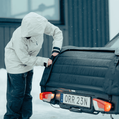 A person wearing a hooded winter coat secures the Thule Onto 2 cargo bag onto the towbar of a car ensuring a firm fit in a cold, snowy setting.
